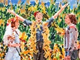 A central figure standing on a pedestal with arms outstretched above him, looking up with joy. Two figures stand on either side of him, and in the background is a lush field of corn. 