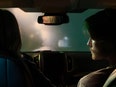 A person turns toward the driver from the passenger seat of a car, while the driver can be seen in the rearview mirror. 